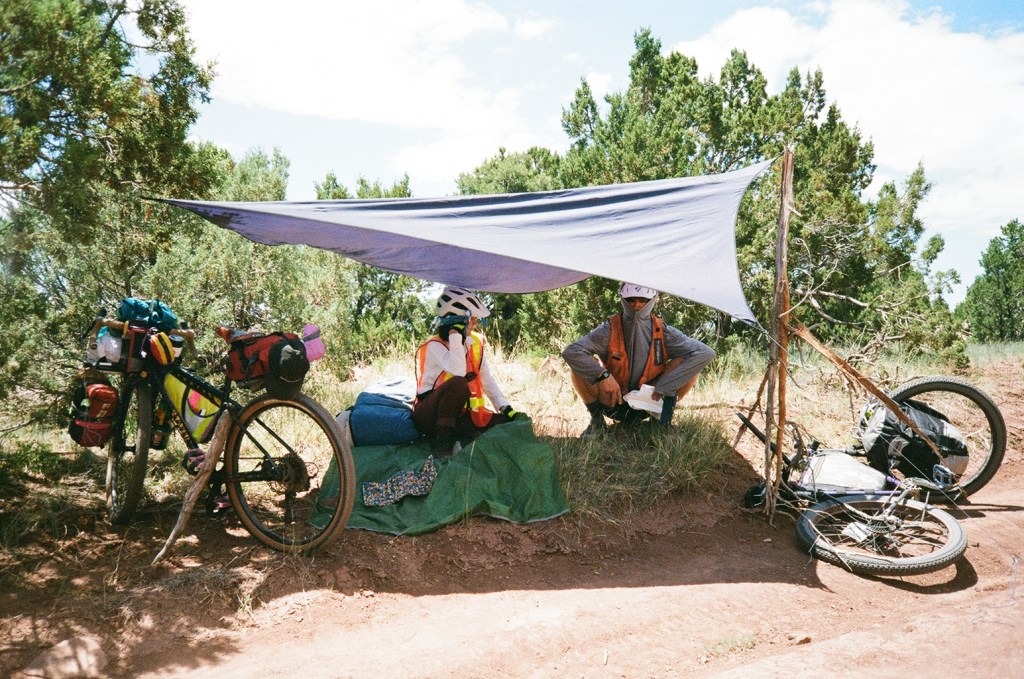 Two cyclists resting under a makeshift tarp shelter in a natural outdoor setting, with bicycles parked beside them.